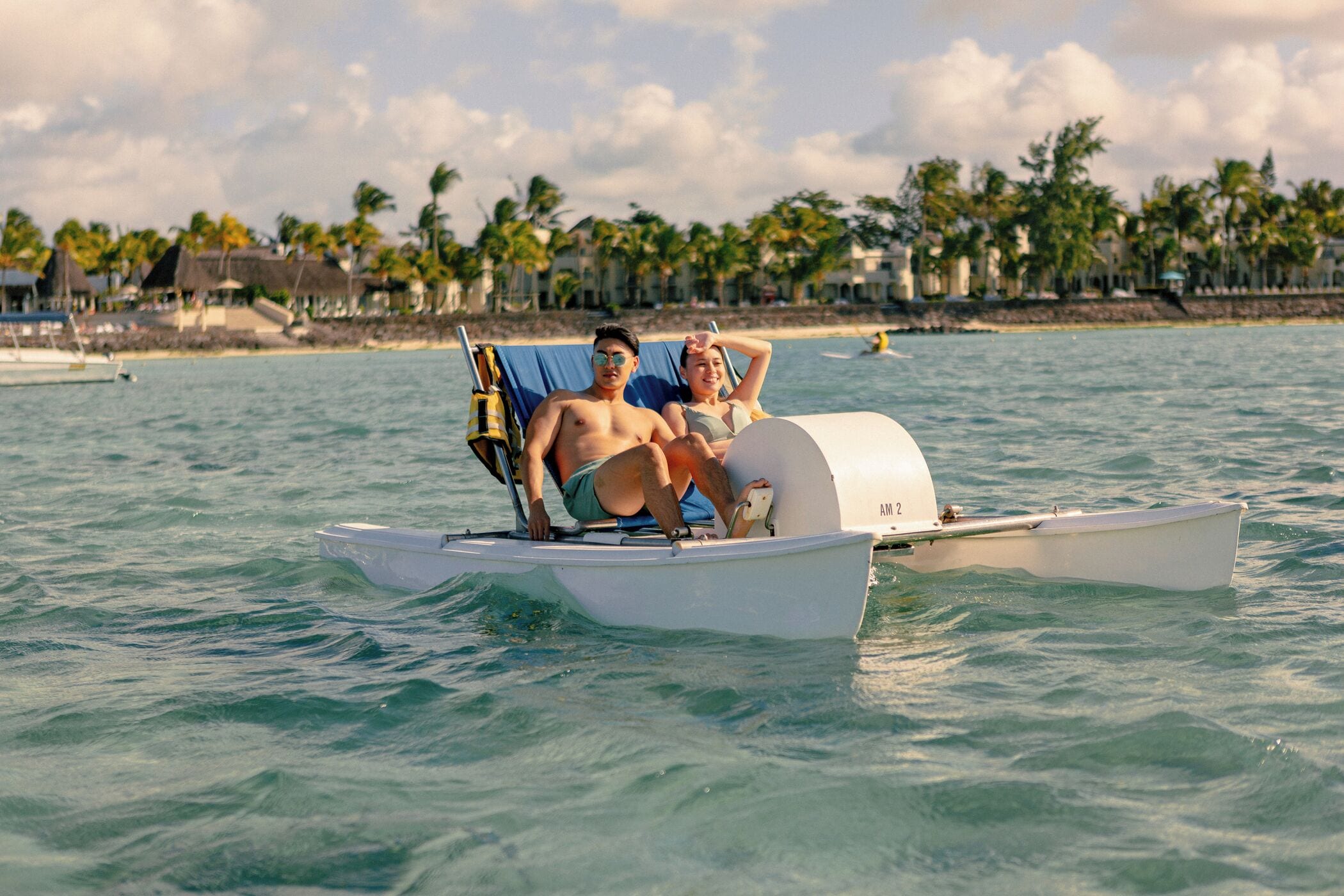 Una pareja en un barco pequeño en un entorno tropical, con palmeras y casas de playa al fondo.