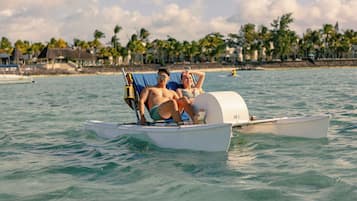 Una pareja en un barco pequeño en un entorno tropical, con palmeras y casas de playa al fondo.