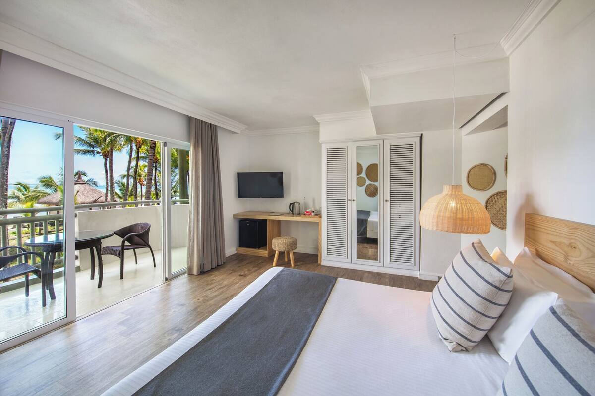 A hotel room with a bed, a desk, a TV, and a balcony with a view of palm trees.