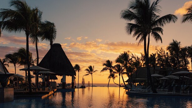 A resort pool area with lounge chairs and umbrellas, palm trees, and a thatched-roof structure. - Ambre - Adults only (Belle Mare)