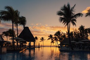 Zona de piscina en un resort, con sillones de descanso y sombrillas, palmeras y una estructura de techo de paja.