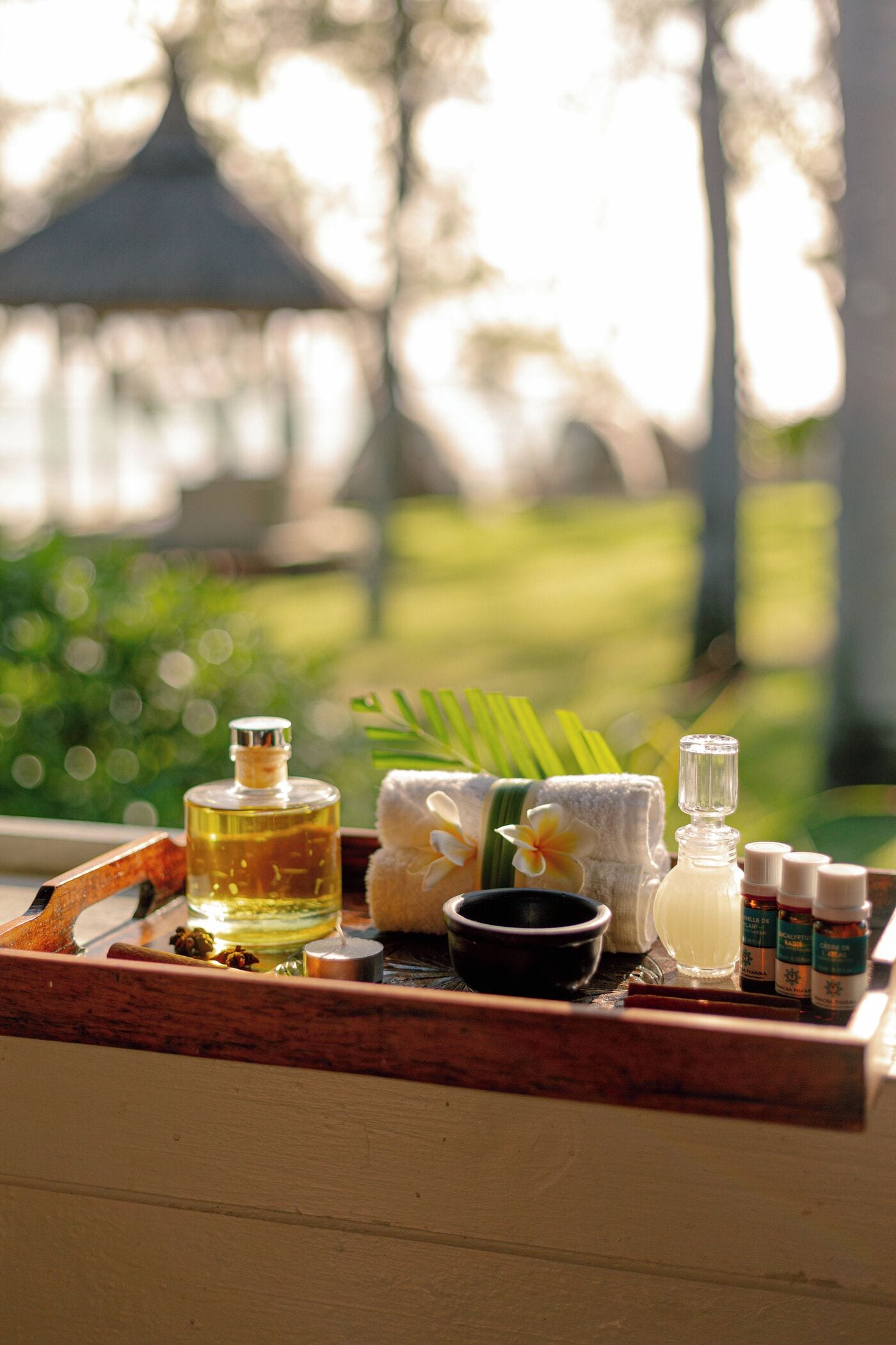 A tray with various spa products, including a bottle of oil, a bowl, and several bottles of essential oils, set against a blurred outdoor background.