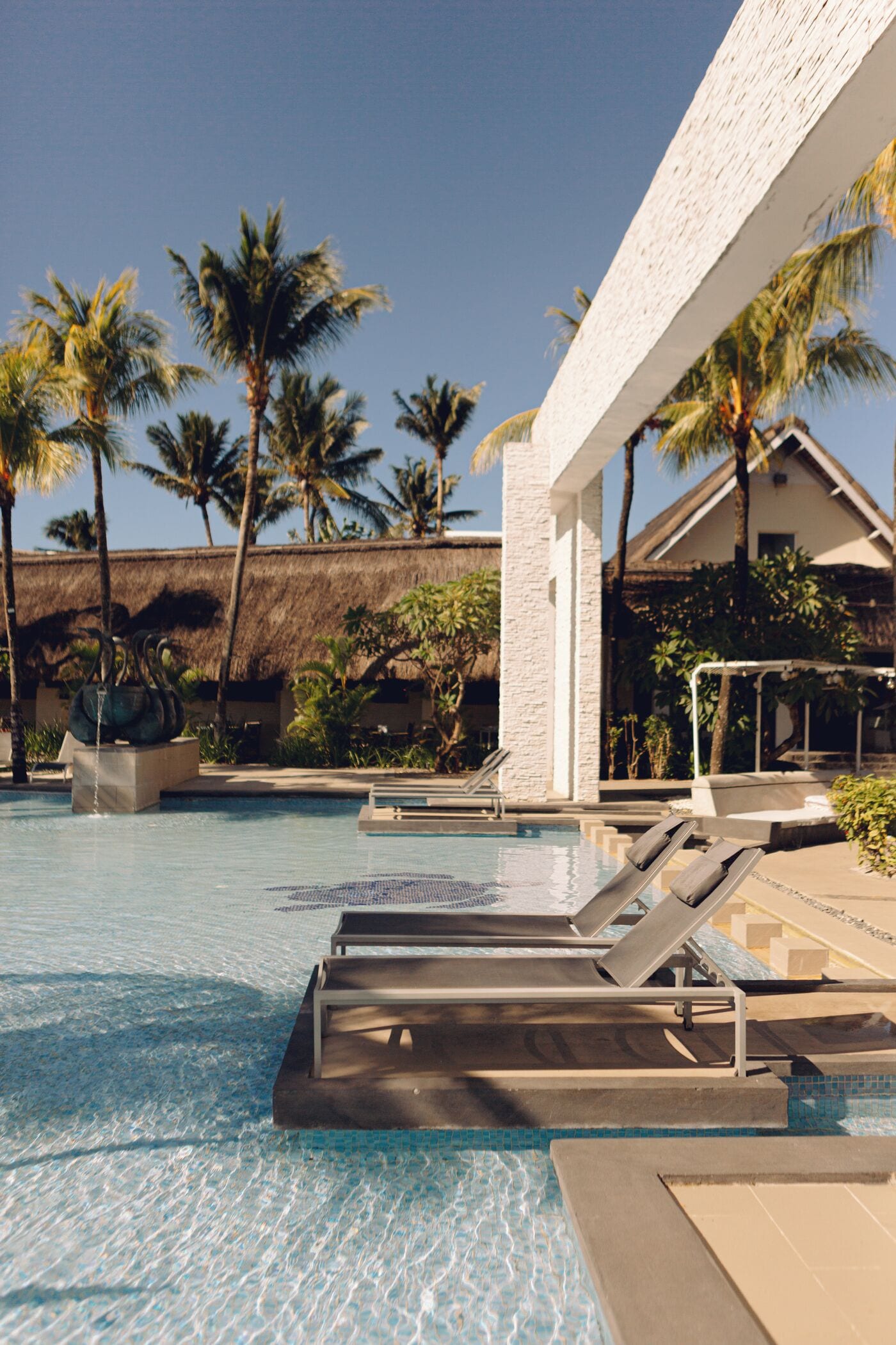 A pool area with lounge chairs, palm trees, and a thatched-roof building.