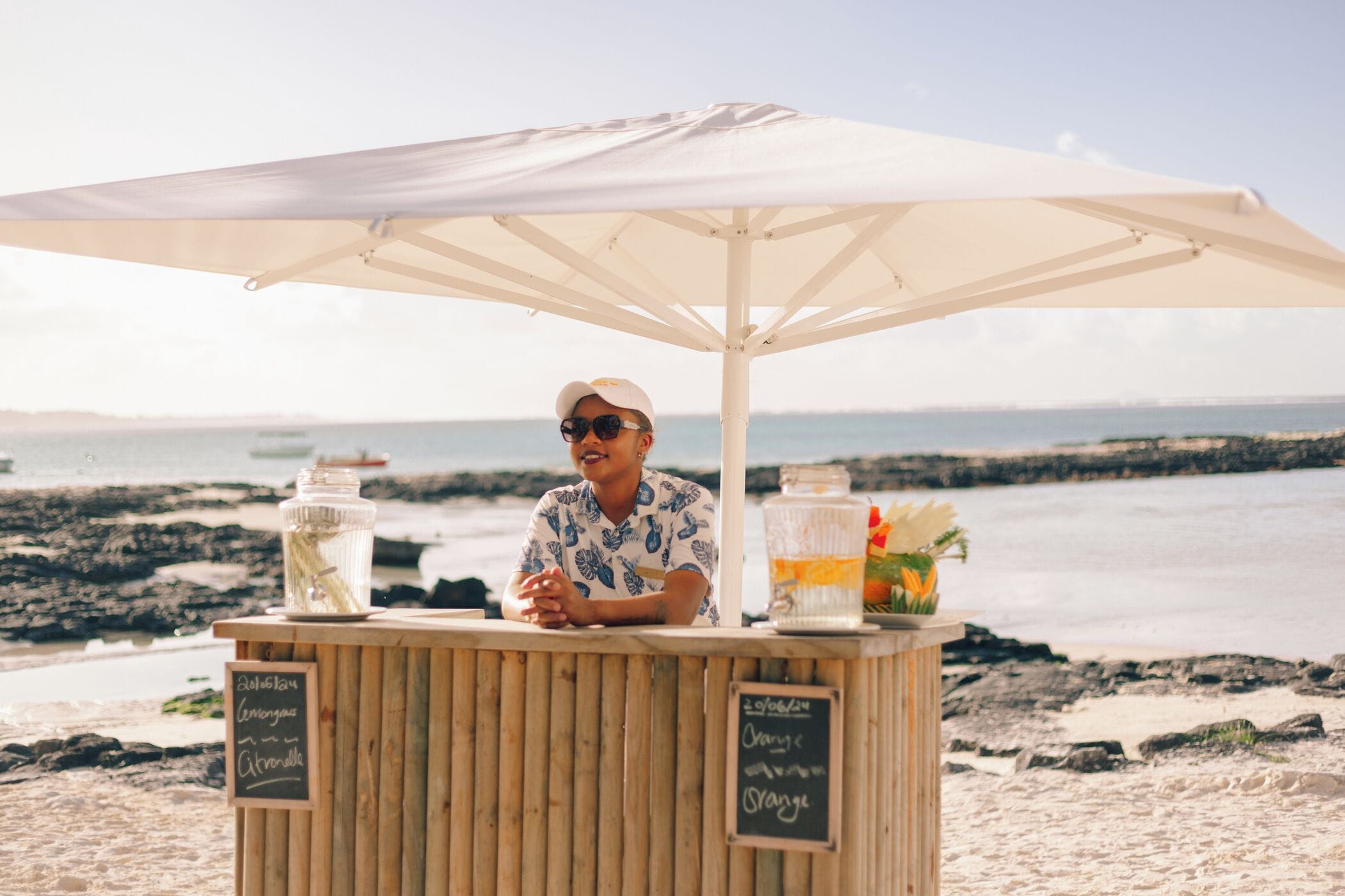A wooden kiosk with a white umbrella on a beach with rocky shoreline and ocean in the background.