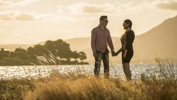 A couple holding hands in a field with a body of water and mountains in the background.