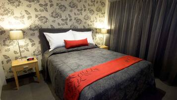 A neatly made bed with a grey and red blanket, two bedside lamps, and a floral wallpaper pattern.