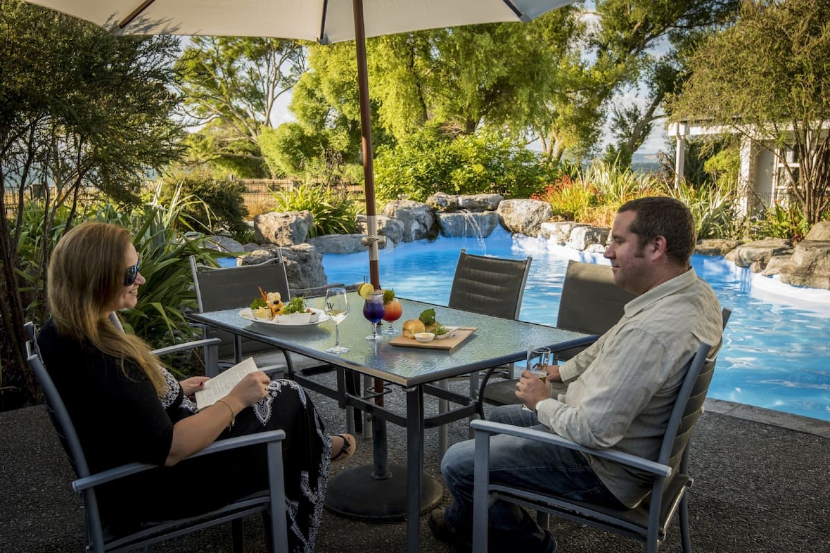 Two people seated at an outdoor table with a view of a pool and waterfall.