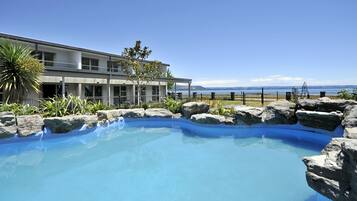 A swimming pool with a rocky edge, surrounded by a building with large windows and a view of the sea.