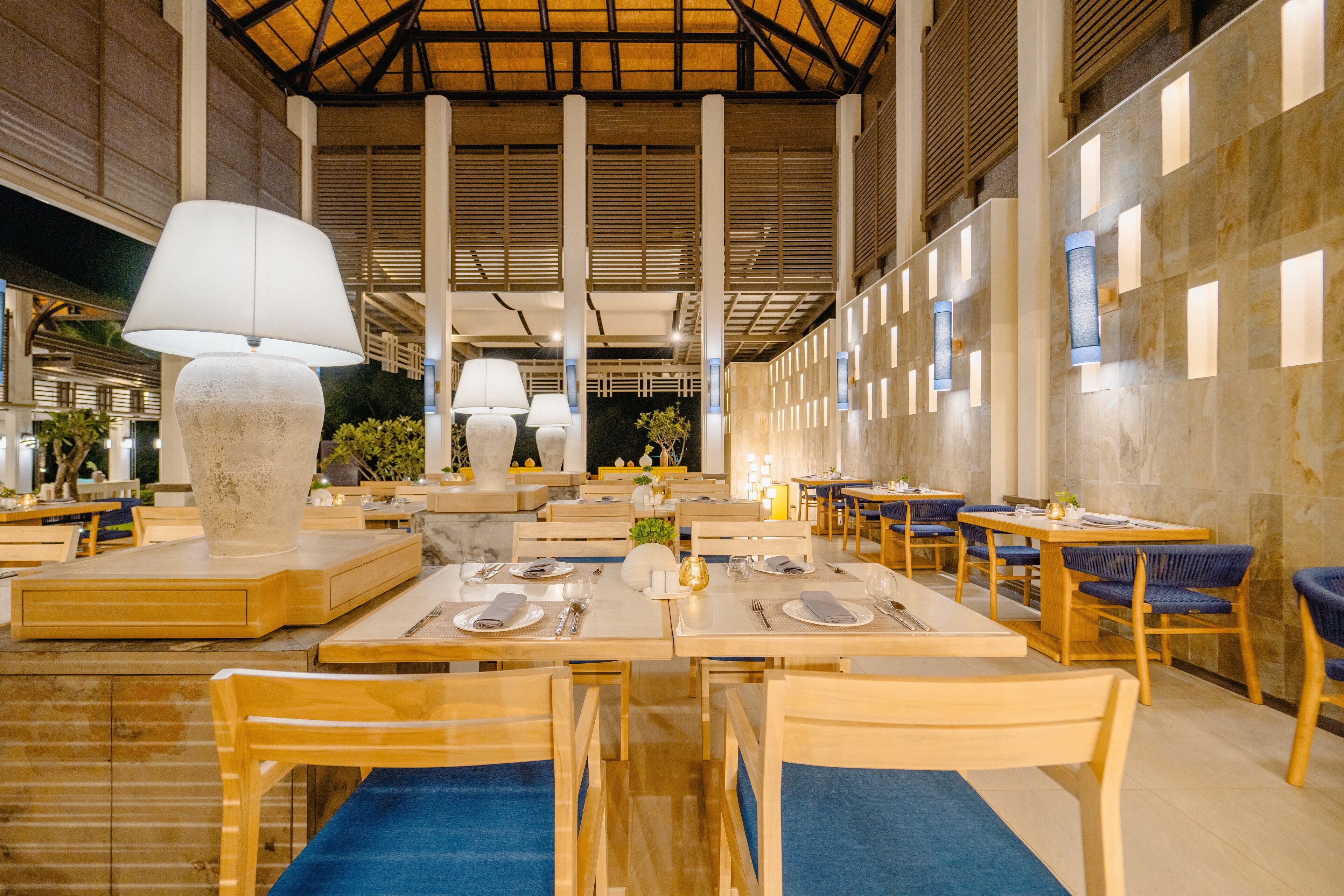 A spacious restaurant with high ceilings, wooden tables, and chairs, and large floor lamps.