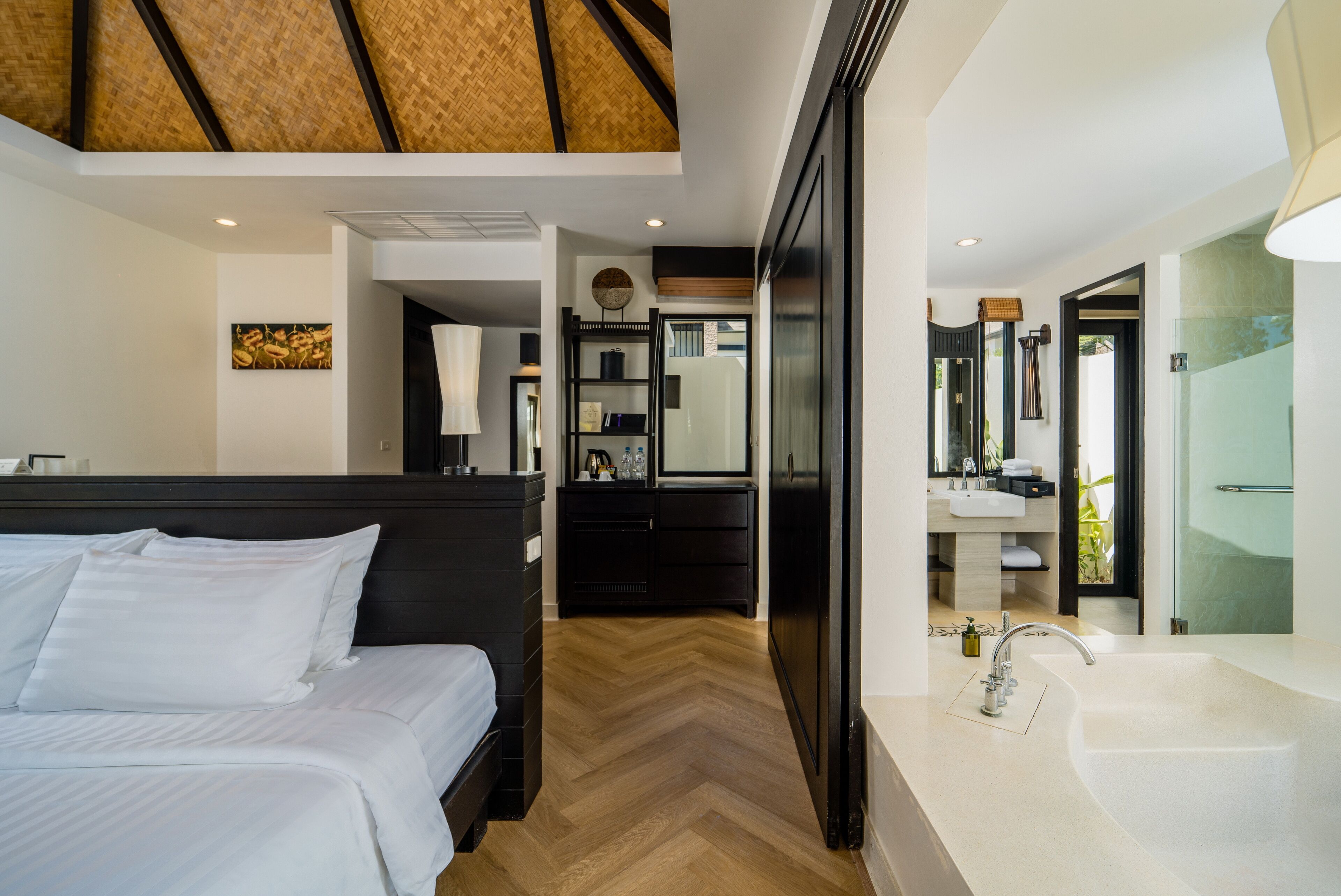 Una habitación de hotel moderna con una cama grande, baño separado con lavamanos y espejo amplios, y piso de madera con patrón de espiga.