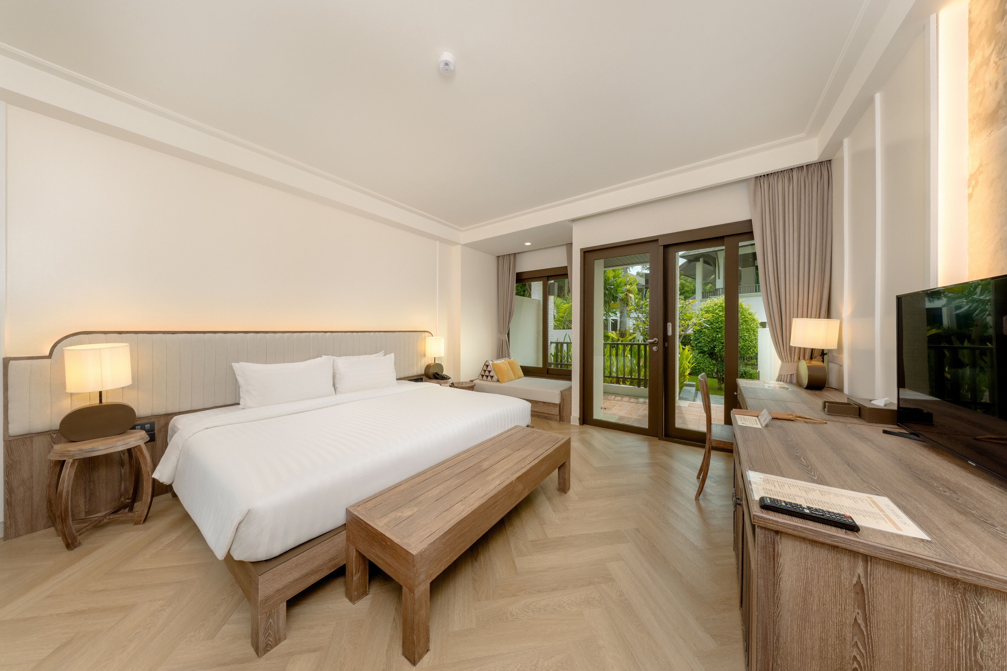 A hotel room with a large bed, a wooden bench, a desk with a TV, and a sliding glass door to the outside.
