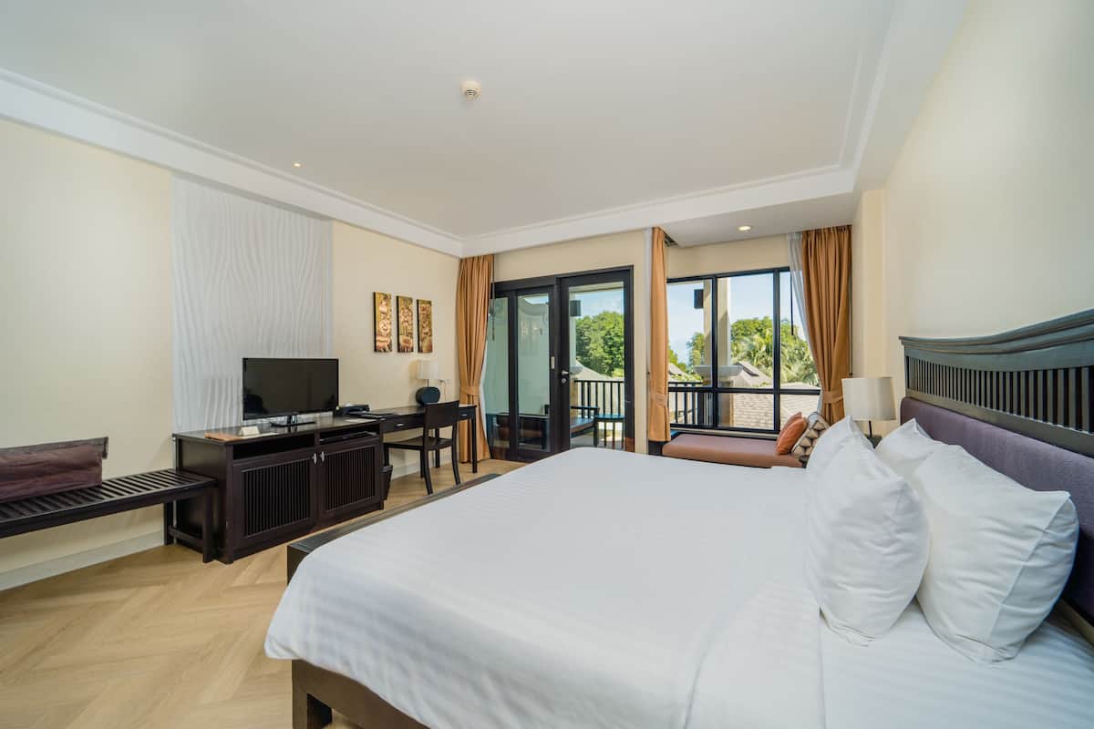 Quarto de hotel com uma cama grande, uma escrivaninha com computador, televisão e varanda com vista para o verde.
