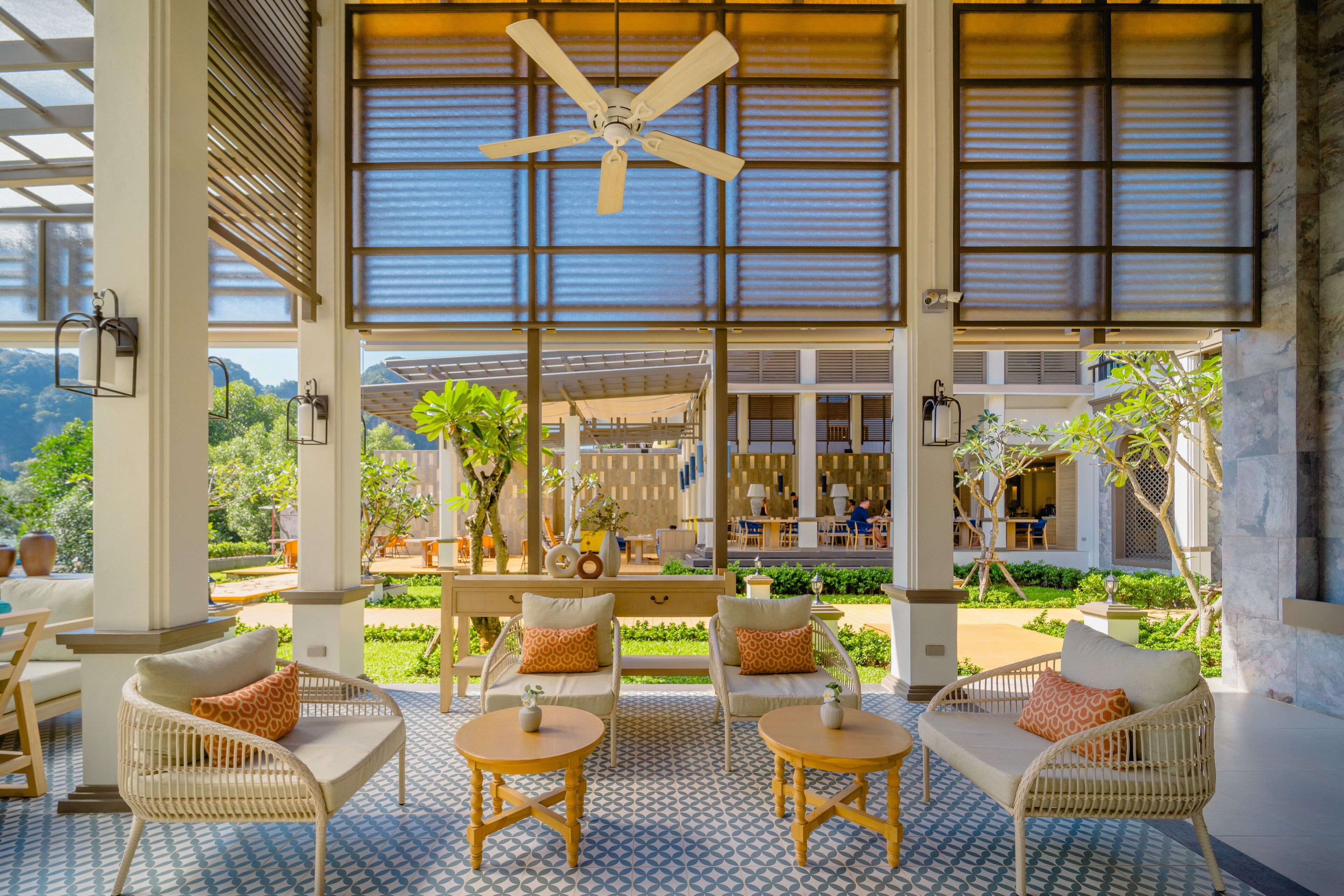An outdoor seating area with wicker furniture and a patterned floor, overlooking a lush garden and a building with large windows.
