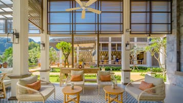 An outdoor seating area with wicker furniture and a patterned floor, overlooking a lush garden and a building with large windows.