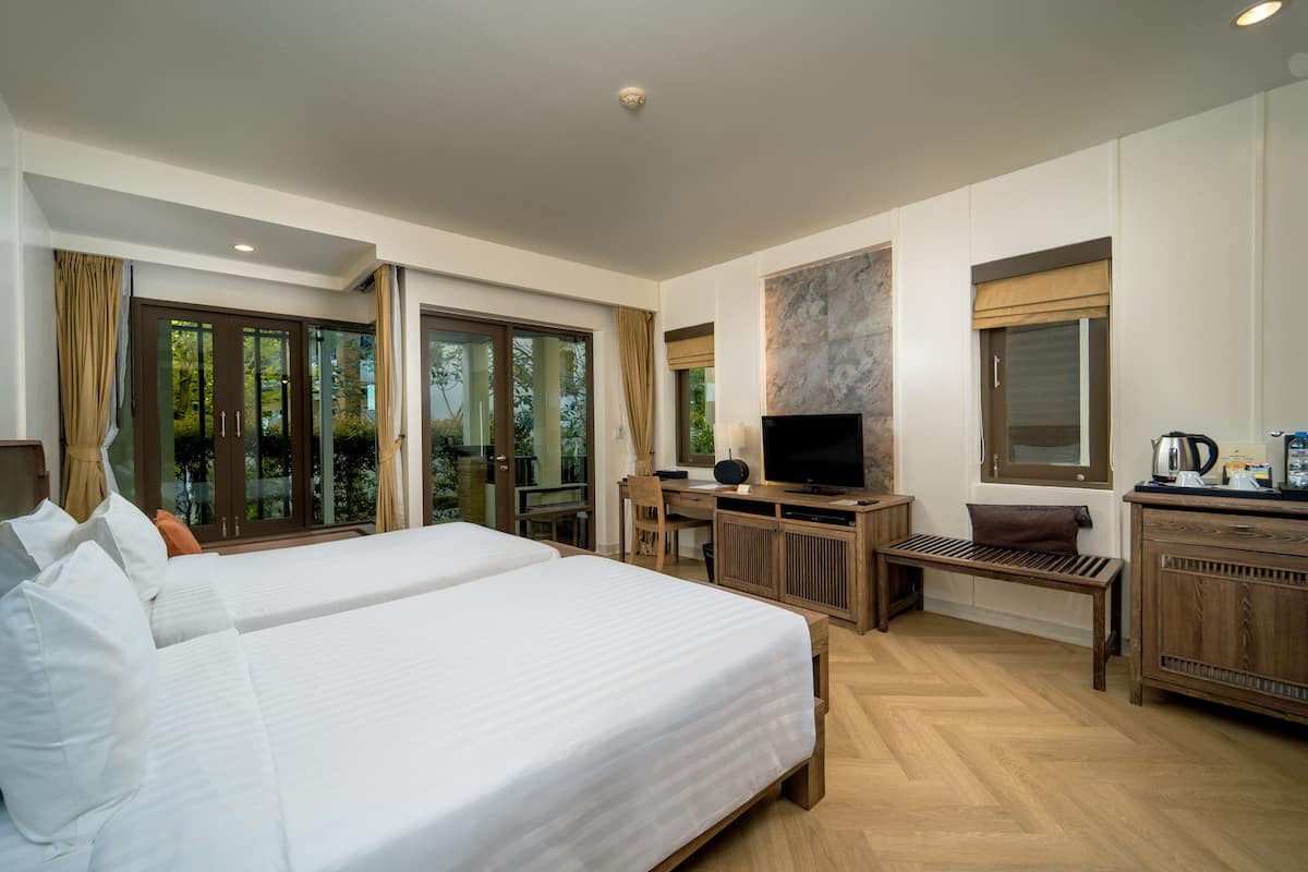 Quarto de hotel com duas camas, uma mesa de trabalho com televisão e vista para o verde através de grandes janelas.