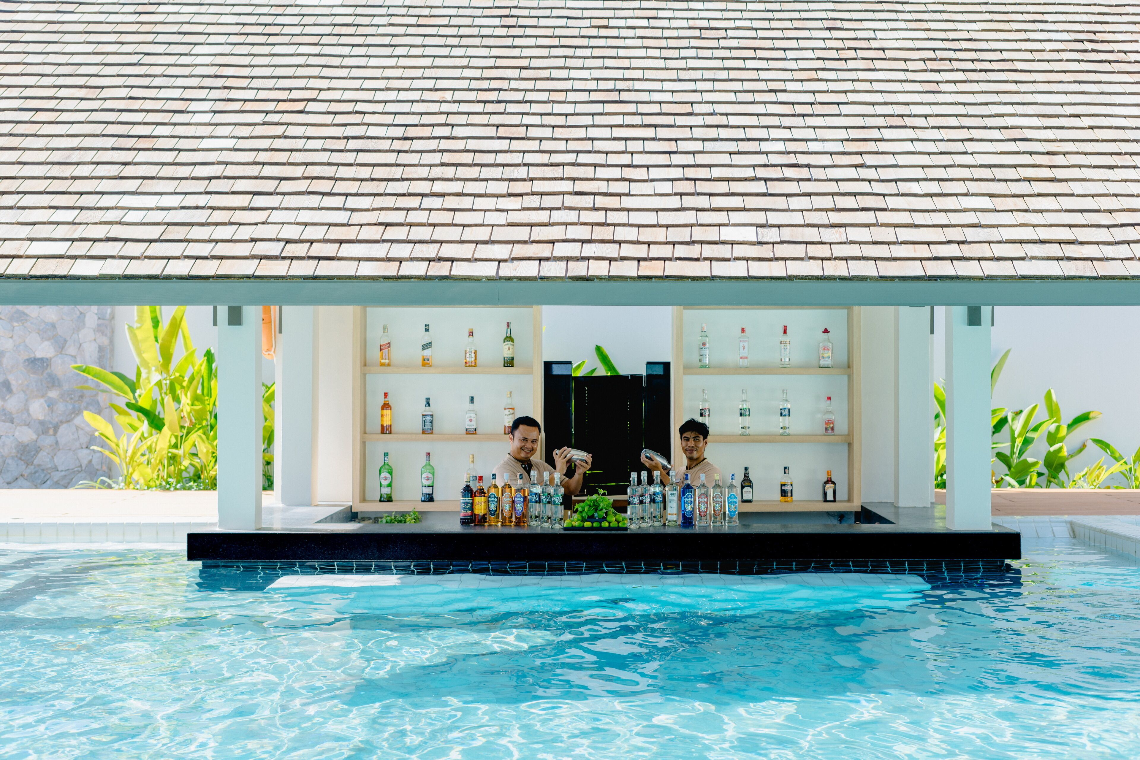 A poolside bar with a tiled roof, shelves of bottles, and a pool.