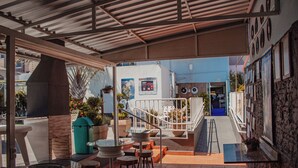 Un espace extérieur aménagé en terrasse, avec des tables et des tabourets, un escalier menant à un niveau supérieur et une vue sur la rue.