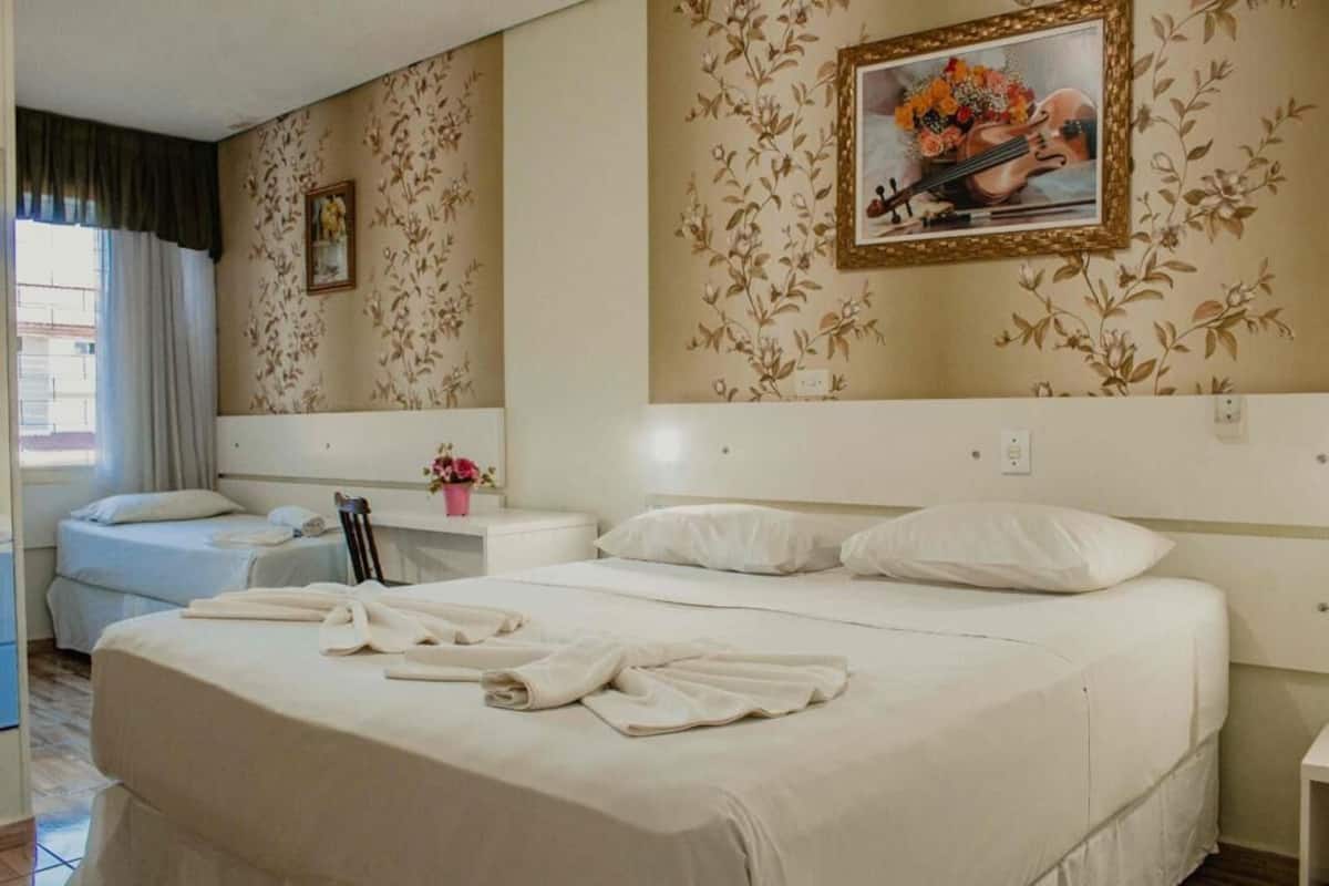 Une chambre d’hôtel avec un grand lit, un lit plus petit, une table de chevet avec une lampe, un tableau au mur et une fenêtre avec des rideaux.
