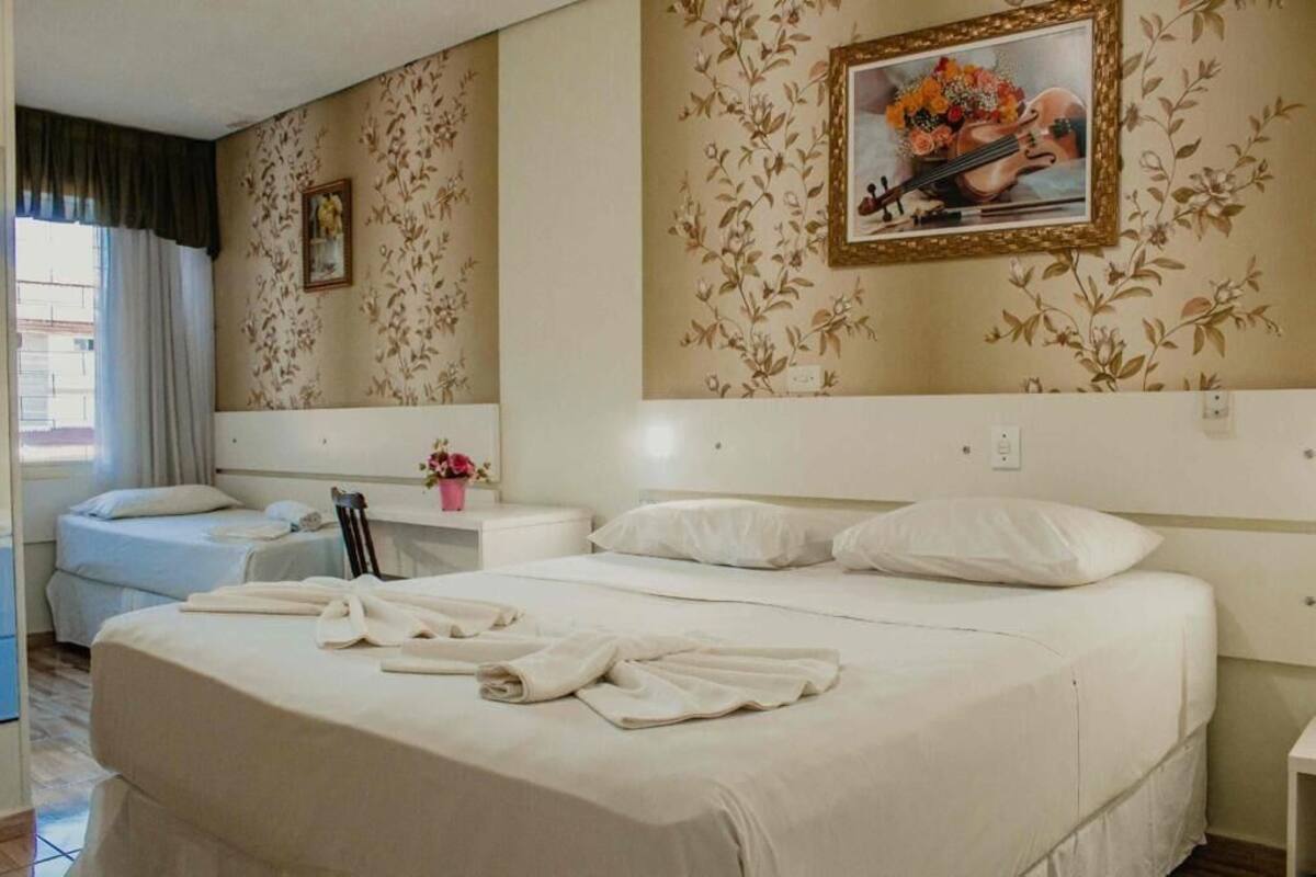 Une chambre d’hôtel avec un grand lit, deux tables de chevet, une commode, une fenêtre avec des rideaux et un papier peint à motifs floraux.