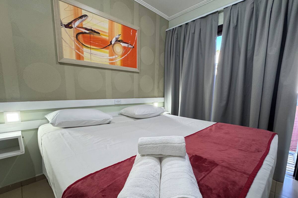 Une chambre d’hôtel avec un lit, des oreillers blancs, une couverture rouge et blanche, et un tableau accroché au mur.