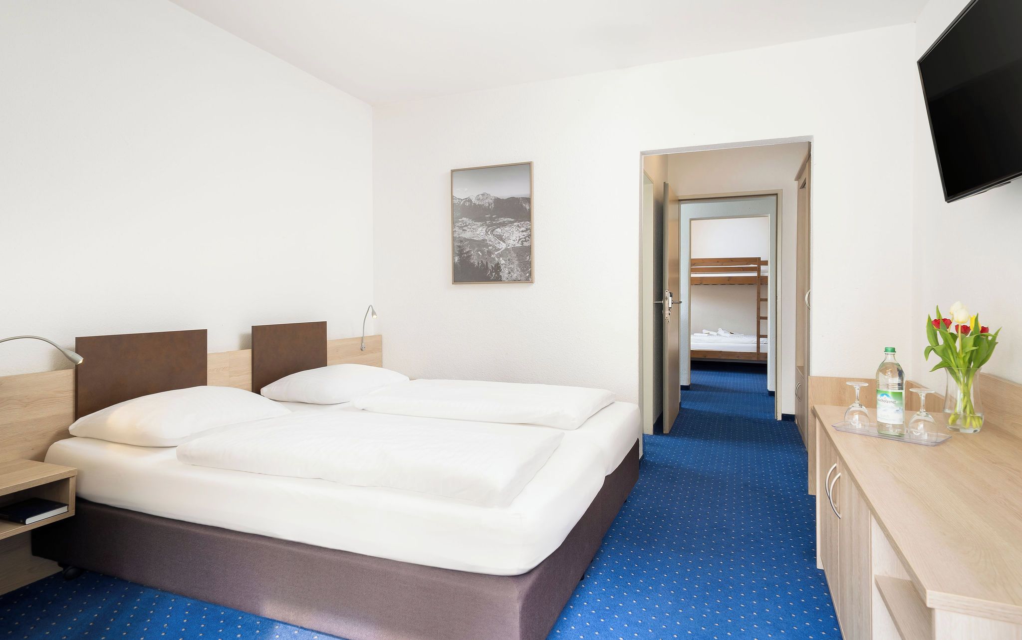 A hotel room with two beds, a desk with a bottle and glasses, a TV, and a picture on the wall.