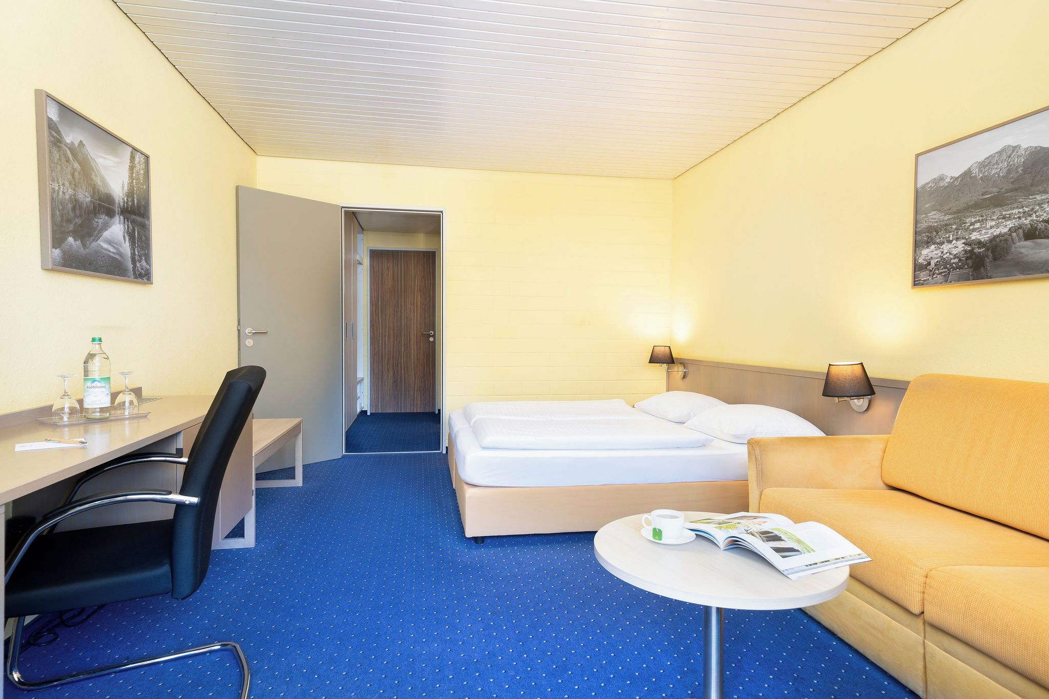 Quarto de hotel com cama, escrivaninha, cadeira, mesinha de centro com xícara e revista, e duas luminárias na parede.
