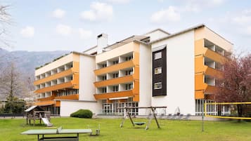A multi-story building with a flat roof, a playground with swings and a slide, and a tennis court.