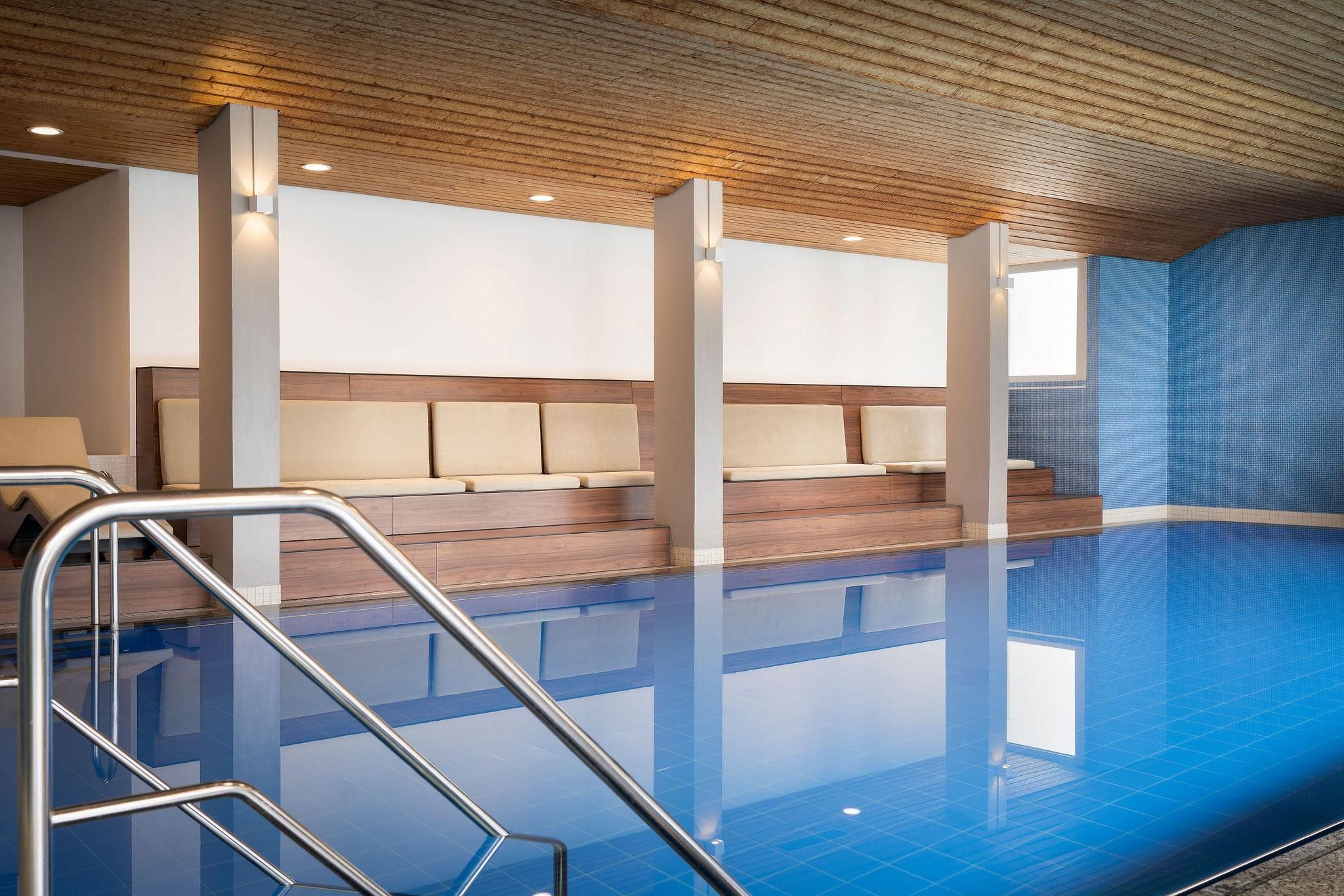 Una piscina con techo de madera, un área de descanso con cojines y una pared de azulejos.