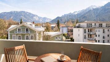 Ein Balkon mit zwei Holzstühlen und einem runden Tisch, mit Blick auf eine Stadt, Gebäude und Berge im Hintergrund.