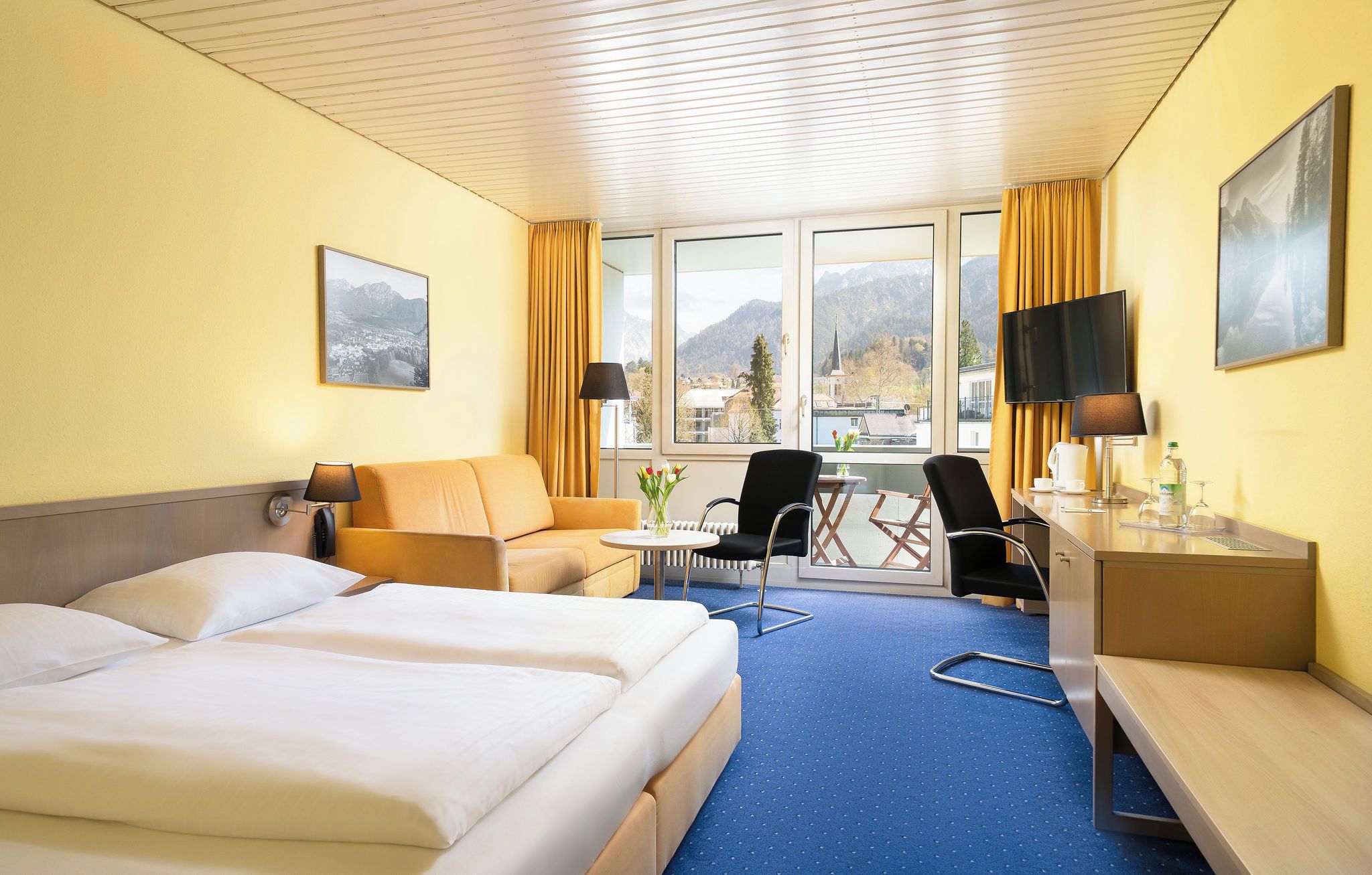 Ein Hotelzimmer mit Bett, Sofa, Schreibtisch und Stuhl. Es gibt Fenster mit Vorhängen, einen Fernseher und ein Bild an der Wand.