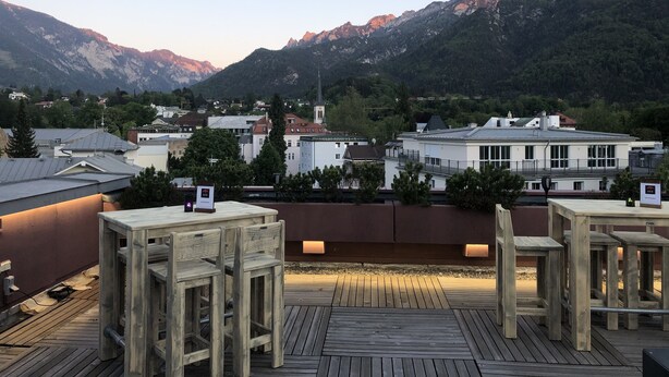 A wooden deck with seating, overlooking a town with buildings and mountains in the background. - Hotel Bayern Vital (Bad Reichenhall)