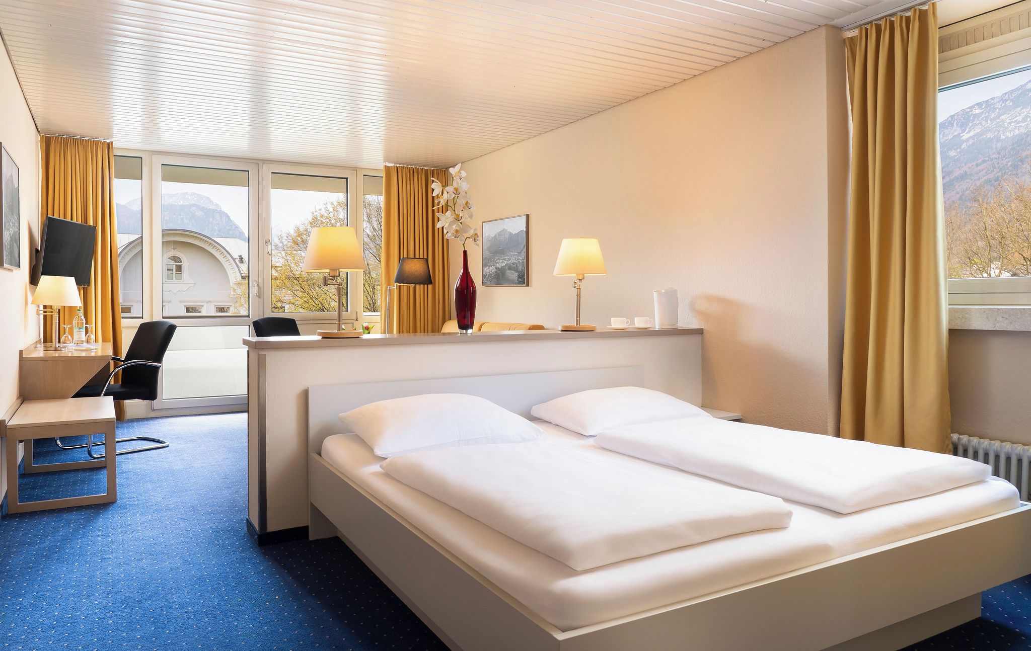 A hotel room with a bed, desk, chair, lamps, and a window with curtains.