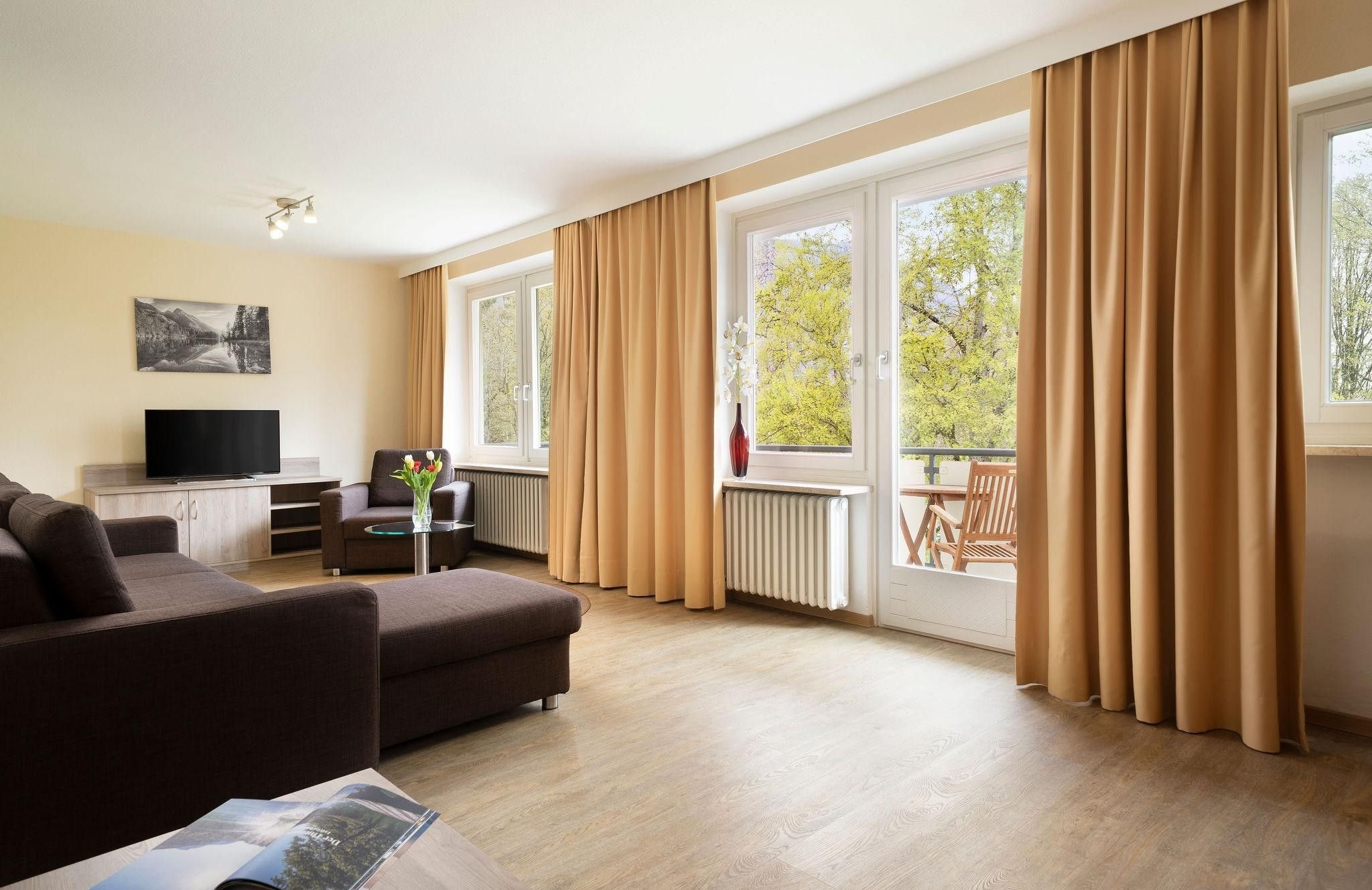 Ein modernes Wohnzimmer mit einer Couch, einem Fernseher, einem kleinen Tisch mit einer Vase und einem Balkon mit Vorhängen.