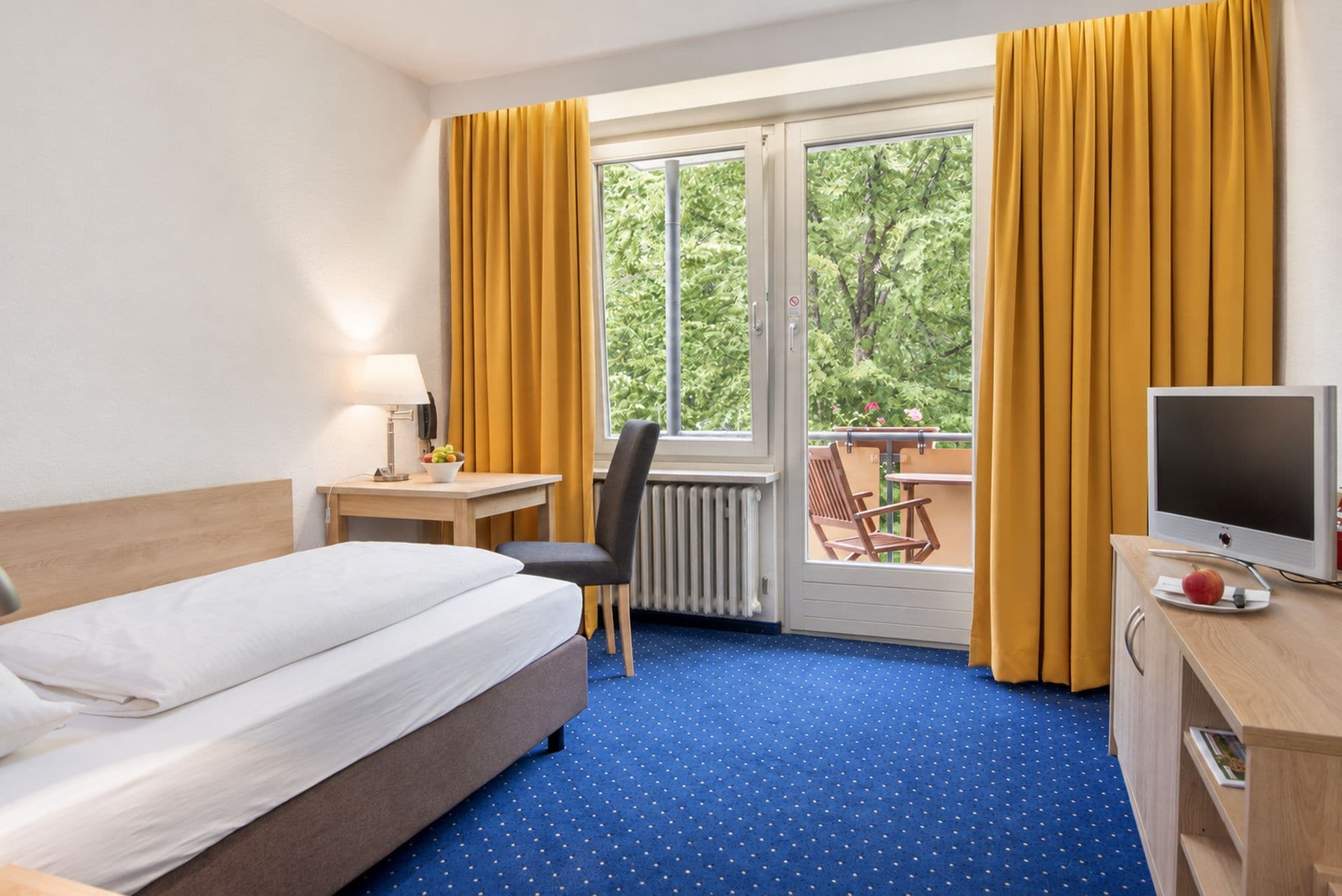 Habitación de hotel con cama, escritorio con computadora, silla, ventana con cortinas y vista a zonas verdes.