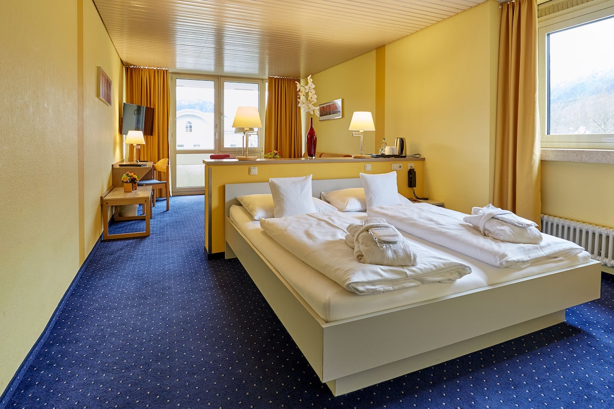A hotel room with a large bed, two bedside lamps, a desk with a chair, a TV, and a window with curtains.