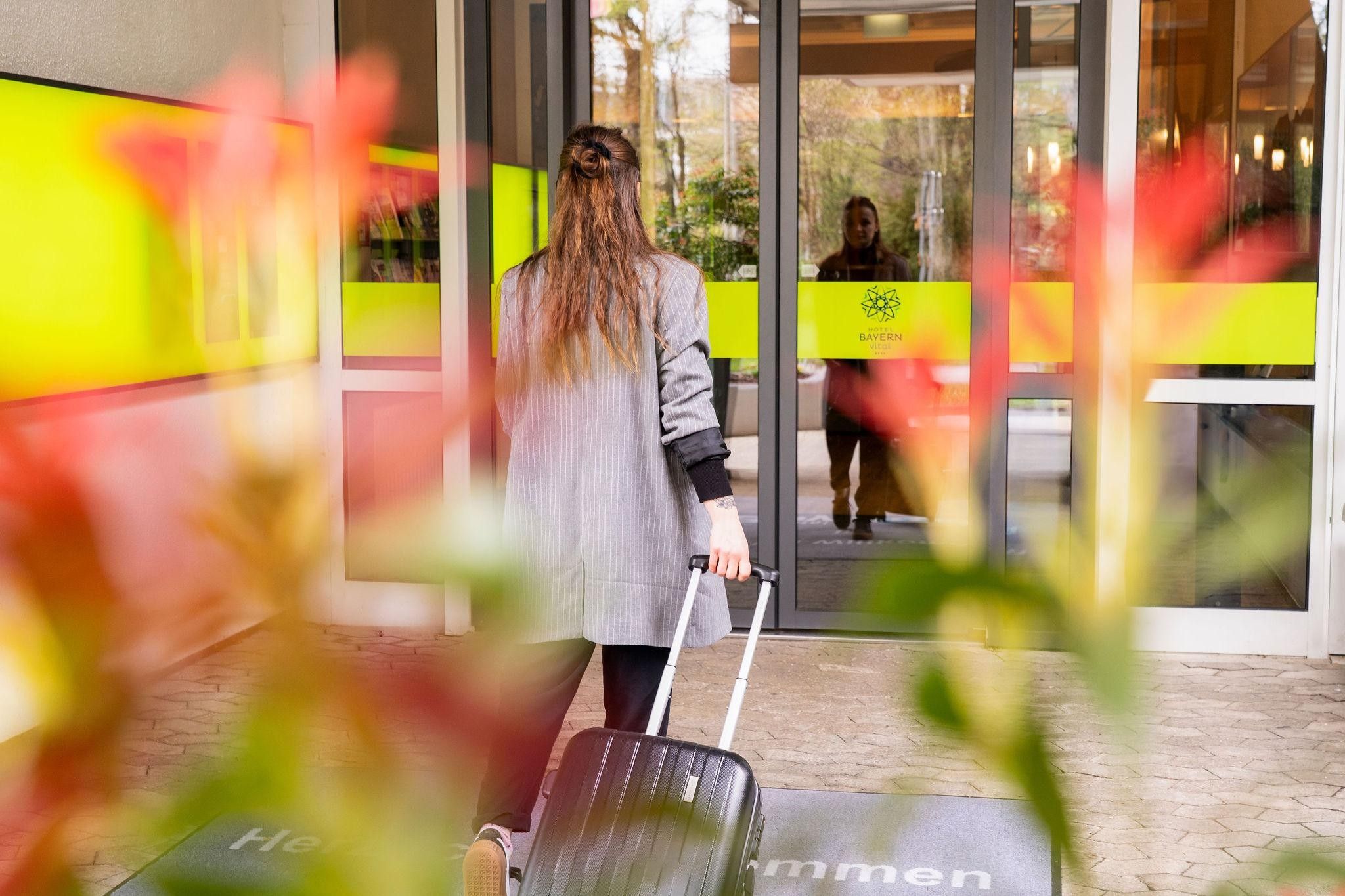 Eine Person mit Koffer betritt ein Gebäude mit Glastür, erkennbarem Logo und Schild mit dem Namen des Betriebs.