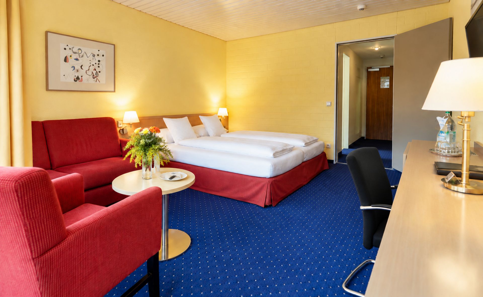 Ein Hotelzimmer mit einem Bett, einem roten Sofa, einem kleinen runden Tisch mit Blumenvase, einem Schreibtisch mit Lampe und einem Wandbild.