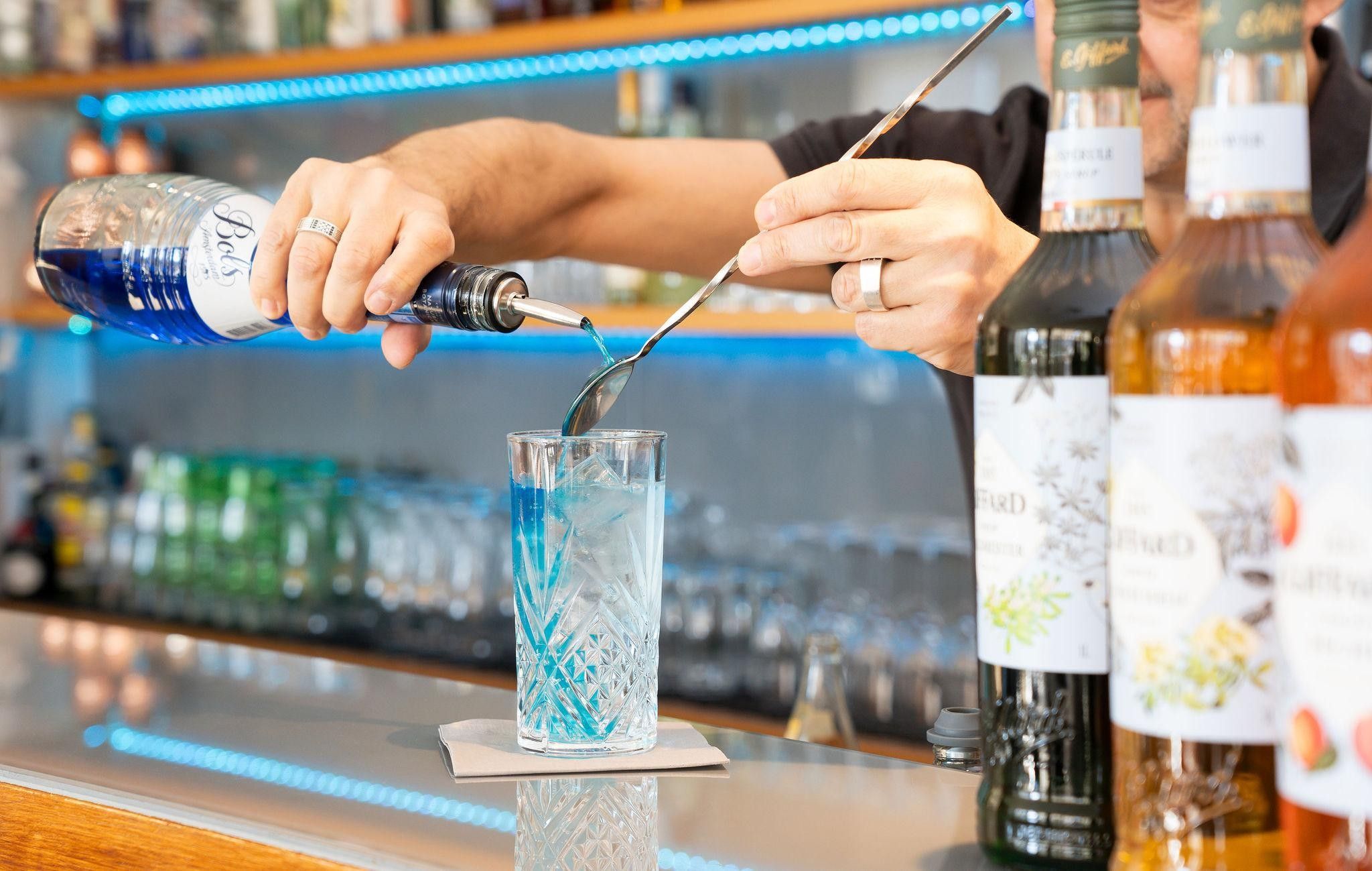 Een bartender giet een blauwe vloeistof in een glas met een metalen hulpmiddel.