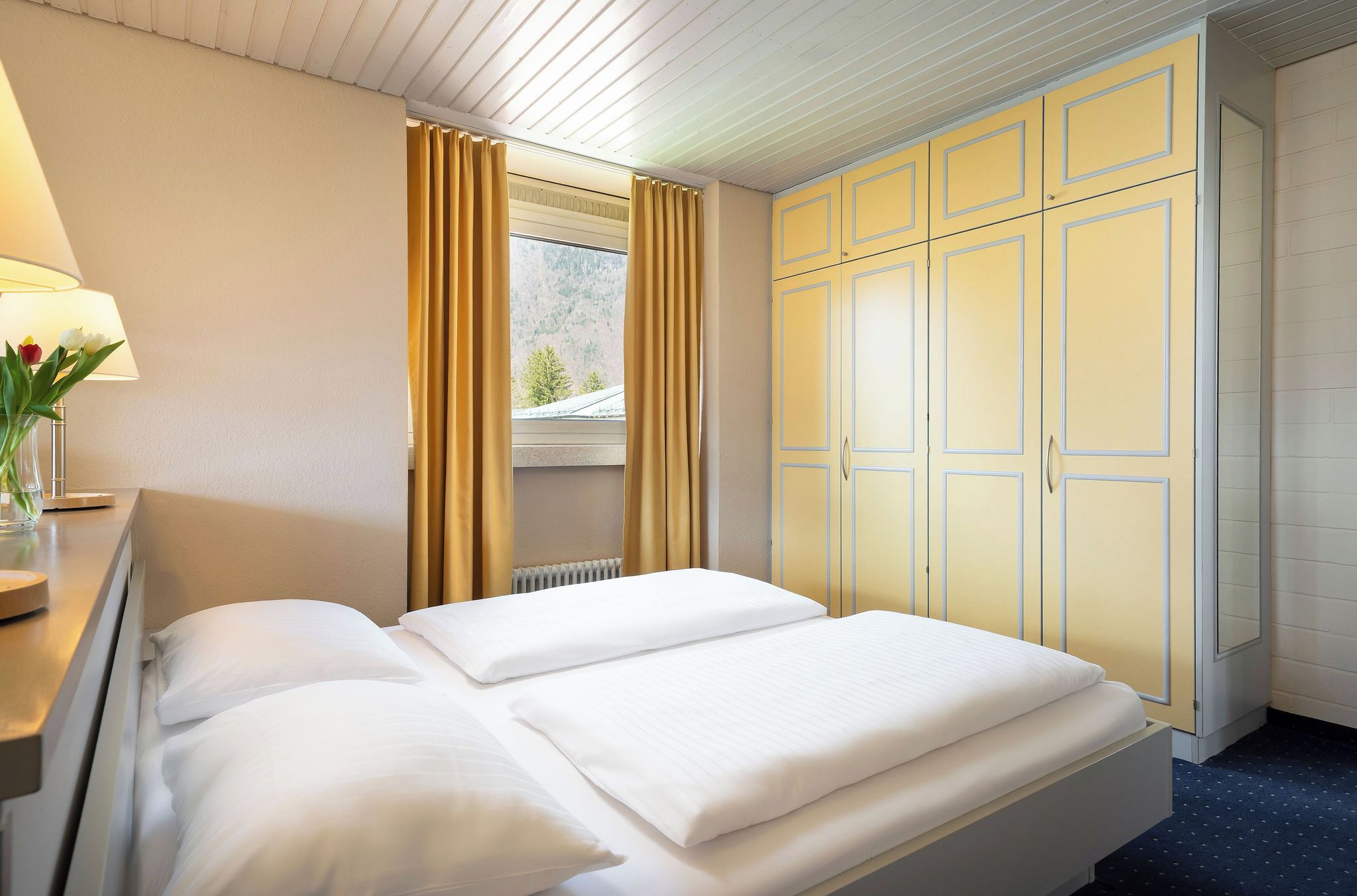 Une chambre d’hôtel comprenant un lit, une table de chevet, une lampe, une fenêtre avec des rideaux et une armoire.