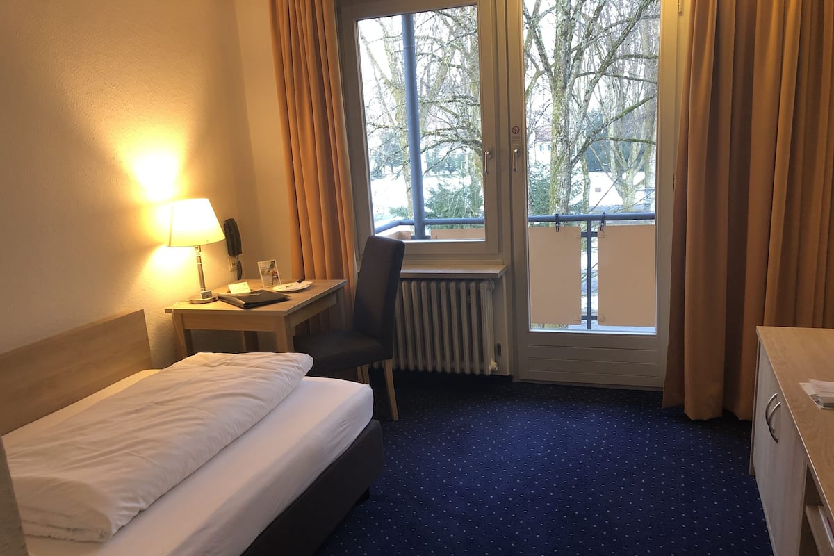 A hotel room with a bed, a desk with a lamp, a chair, a radiator, and a window with curtains.