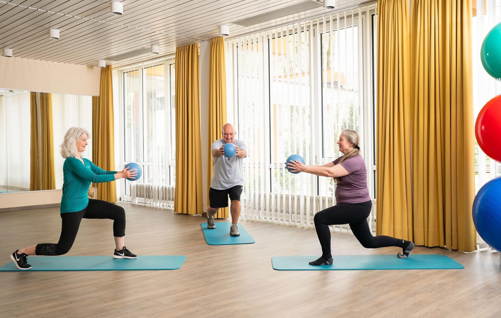 Een fitnessles met deelnemers op matten, gebruikmakend van fitnessballen, in een ruimte met grote ramen en gordijnen.