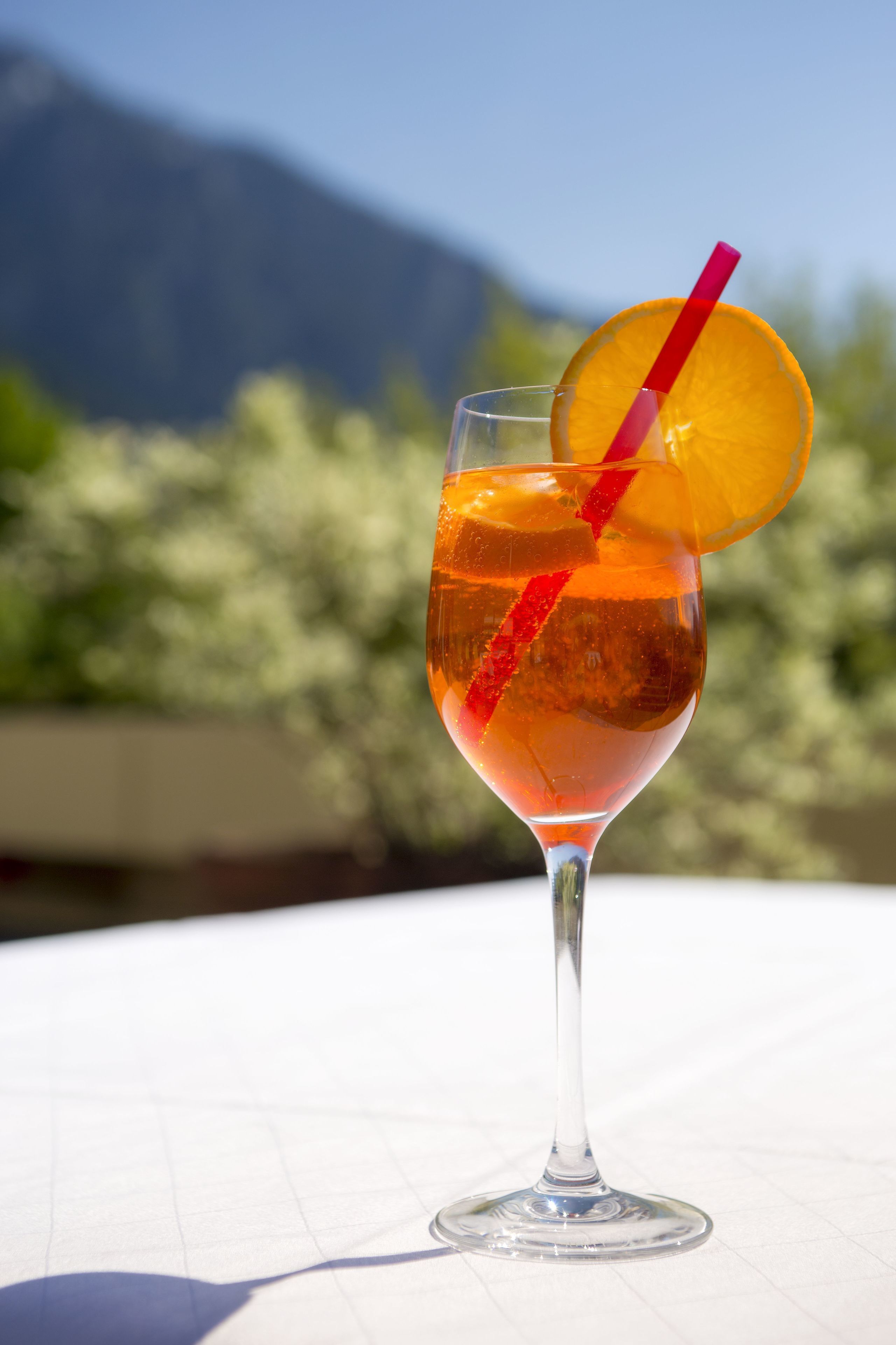 Un vaso de cóctel con una rodaja de naranja y una pajita roja sobre una mesa, con montañas al fondo.