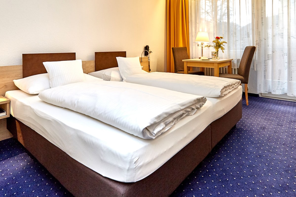 A hotel room with two beds, a desk, a chair, a lamp, and a window with curtains.