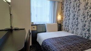 A hotel room with a bed, a desk, a chair, a window with curtains, a wall with a floral pattern, and a bedside table with a lamp. - APA Hotel Okayama Ekimae (Okayama)