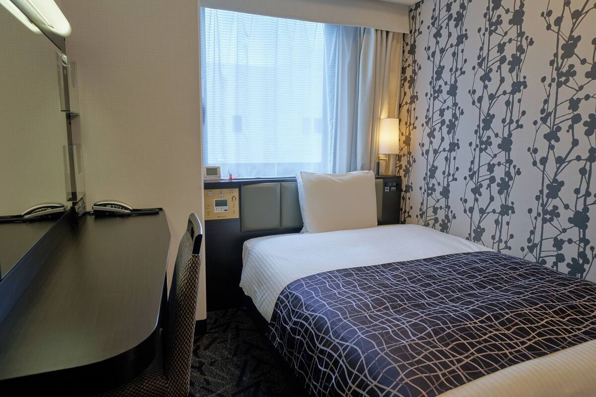 A hotel room with a bed, a desk, a chair, a window with curtains, a wall with a floral pattern, and a bedside table with a lamp.