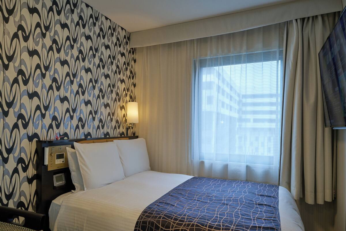 A hotel room with a large bed, a bedside table with a lamp, a wall clock, and a window with curtains.