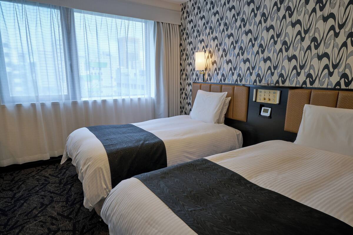 A hotel room with two beds, a patterned wall, a window with curtains, and a wall-mounted control panel.