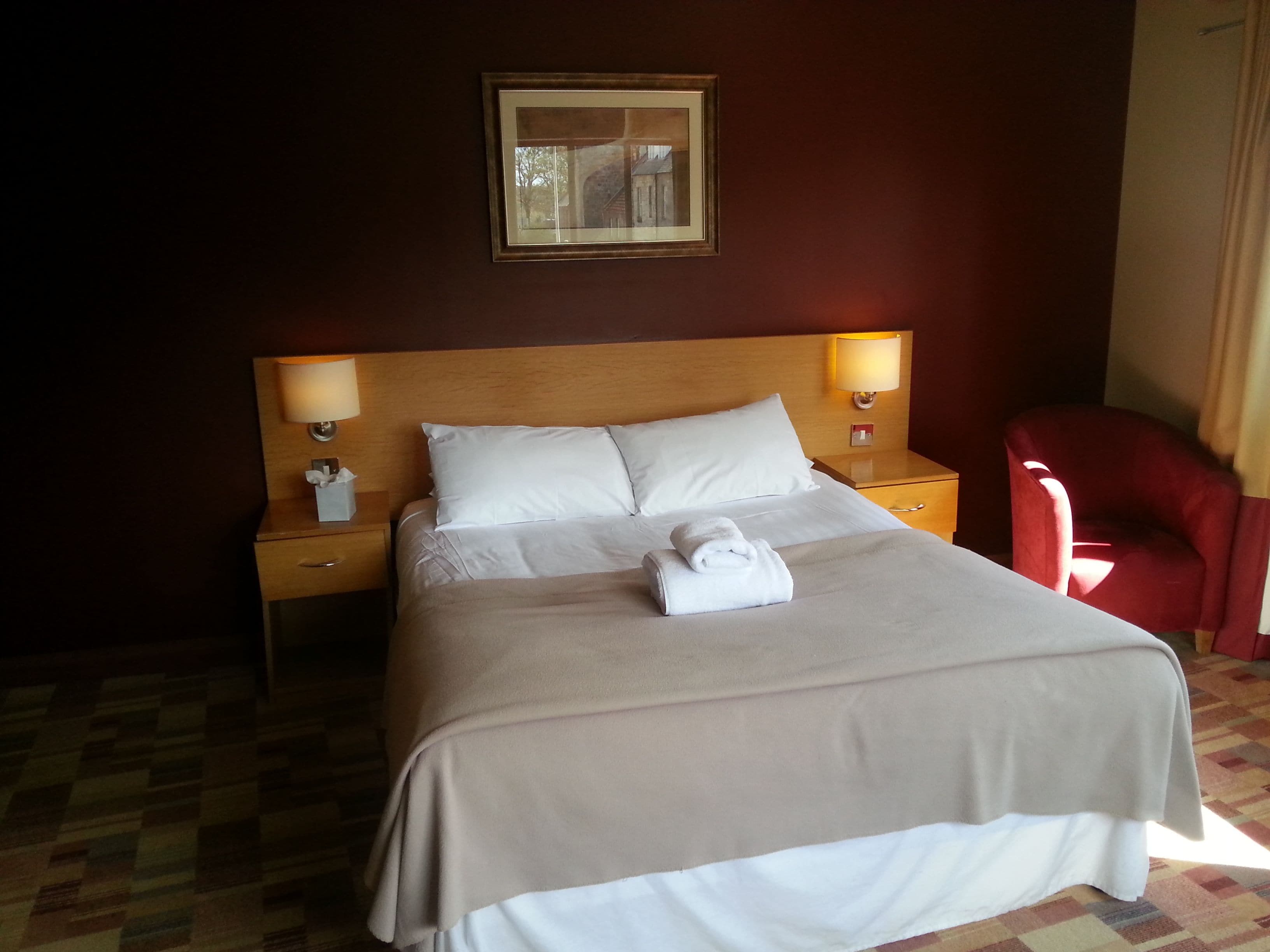 A neatly made bed with a white comforter and pillows, two bedside lamps, a red chair, and a framed picture on the wall.