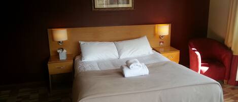 A neatly made bed with a white comforter and pillows, two bedside lamps, a red chair, and a framed picture on the wall.