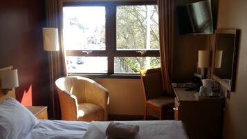 A hotel room with a bed, two chairs, a desk, a TV, and a window with a view of the street.